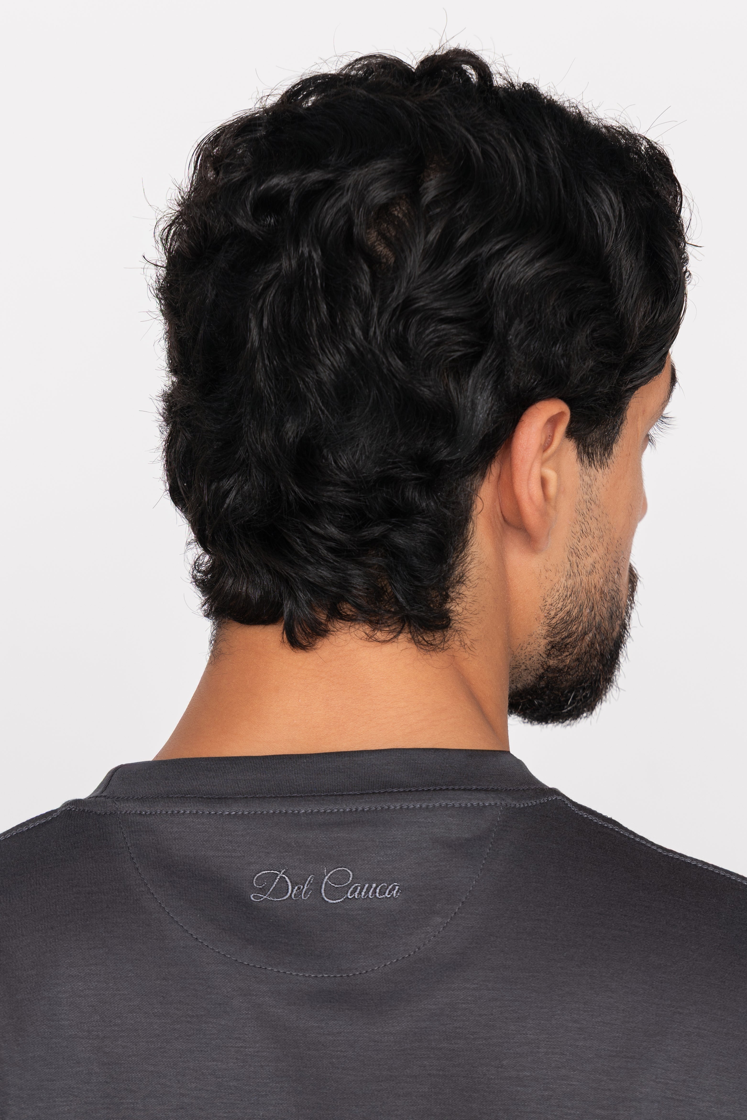 Man with styled hair wearing a dark gray shirt with a brand logo on a light gray background
